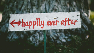 En vit träskylt med röd text som säger "happily ever after" och en pil som pekar åt vänster. Skylten står utomhus framför ett stort träd och förmedlar en känsla av lycka, kärlek och sagoslut.