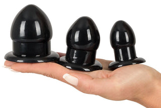 En hand håller tre snygga svarta silikonpluggar från Anal Stretching Plug Kit, perfekt för säker anal stimulering. De har en glansig finish och breda baser, arrangerade från största till minsta för perfekt gradvis träning.