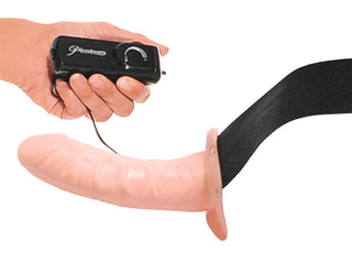 En hand håller i en fjärrkontroll som är fäst vid Vibrating Hollow Strap-On med svart sele, perfekt för pegging-äventyr.