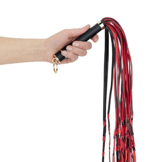 En hand håller i Flogger Piska I PU-Läder 64 cm, en svart och röd flogger i PU-läder med flera svansar och ett handtag med ögla - perfekt för bondagelekar.