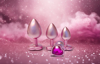 Tre metallpluggar med hjärtformade baser och en Pearl Pink Buttplugg Small vilar på en rosa glittrig yta mot en drömsk, gnistrande bakgrund.