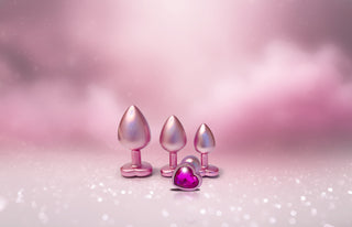 Tre metallicrosa pluggar, inklusive Pearl Pink Buttplugg Small med hjärtformad bas, vilar på en glittrande rosa bakgrund.