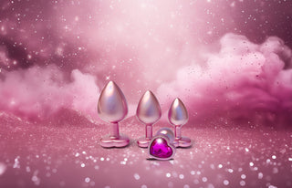Pearl Pink Buttplugg Medium, med en hjärtformad bas och en lila ädelsten, vilar på en gnistrande rosa bakgrund med moln, vilket skapar en lekfull men elegant atmosfär.