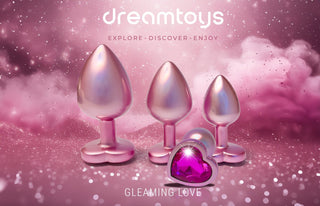 Tre metalliska föremål med en hjärtformad bas har hjärtformade ädelstenar på en rosa, gnistrande bakgrund märkt "drömleksaker" och "Gleaming Love Pearl Pink Buttplug Set.