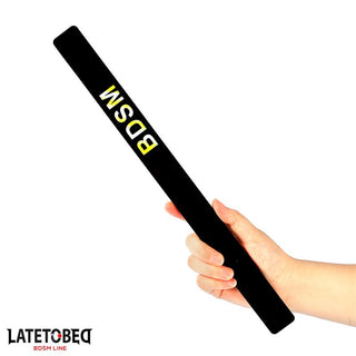 En hand håller i Latetobed Spanking Paddle 38cm, en svart paddel med "BDSM" tryckt i feta gula bokstäver.