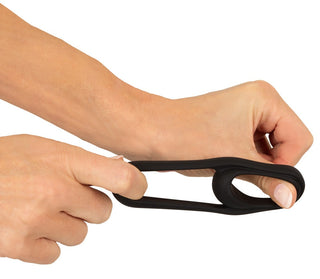 En hand sträcker ut Erecto Cock & Ball Ring, en svart silikonring med en oval öppning, mot en vit bakgrund.