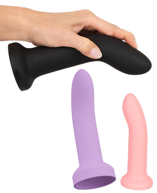 En hand håller en svart dildo från Strap-on Set med 3 Silikondildos över en yta med två andra: en större lila och mindre rosa. Alla tillverkade av flytande silikon, de har släta texturer och utsvängda baser, idealiska för mångsidig användning.