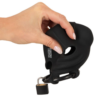 En hand håller en Black Velvets Kyskhetsbur, en svart silikonenhet som liknar ett låsbart föremål med ett centralt hål, gjord av mjukt, flexibelt material.