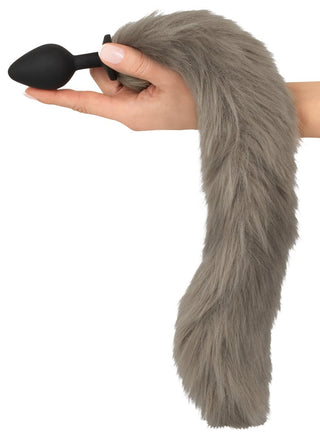 En hand håller Fluffy Tail Buttplug, perfekt för husdjurslek eller cosplay, som visar upp en falsk grå rävsvans fäst vid en elegant svart plugg mot en vit bakgrund.