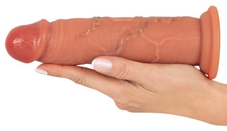 En hand håller Realistixxx Real Skin Dildo, med synliga vener, en utsvängd bas och ett hudtonat, realistiskt utseende mot en vit bakgrund.
