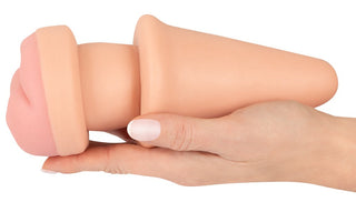En hand håller 2in1 Buttplug Extension, en köttfärgad silikonleksak för vuxna som är utformad för mångsidig njutning.