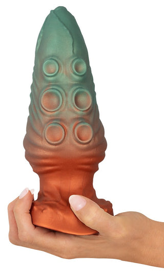 En hand som håller i Beasty Cocks Atlantis Buttplug, en stor, texturerad grön och bronsfärgad analplugg tillverkad av flytande silikon med cirkulära mönster.