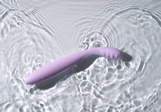 Den lavendelfärgade, böjda Svakom Cici 2 Vibrator flyter på klart vatten och skapar krusningar. Dess strukturerade yta har små vågor för en lugnande effekt. Anslut den till SVAKOM-appen för en flexibel och anpassningsbar upplevelse.