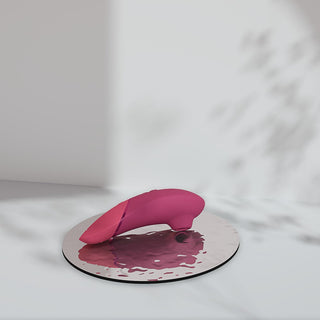 Rosa silikon Womanizer NEXT, abstrakt design, vilande på reflekterande yta med vattendroppar och mjuka skuggor.