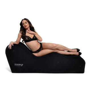 En kvinna i svarta underkläder vilar på Uppblåsbaren The Lounge Sexkudde, en snygg, ergonomiskt designad schäslong. Med långt mörkt hår rör hon vid halsen medan "Bedroom Fantasies"-loggan pryder sidan. Denna bärbara pjäs lyfter alla miljöer med elegans.