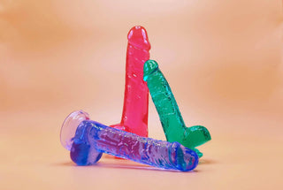 Färgglada genomskinliga dildos i rosa, grönt och blått med realistiska detaljer, placerade mot en mjuk beige bakgrund. Perfekt för vår dildo-kategori