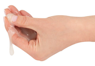 Närbild av en hand som håller genomskinliga Fejk Sperma Ananas, en vattenbaserad vätska, medan den försiktigt rinner genom fingrarna mot en vit bakgrund.
